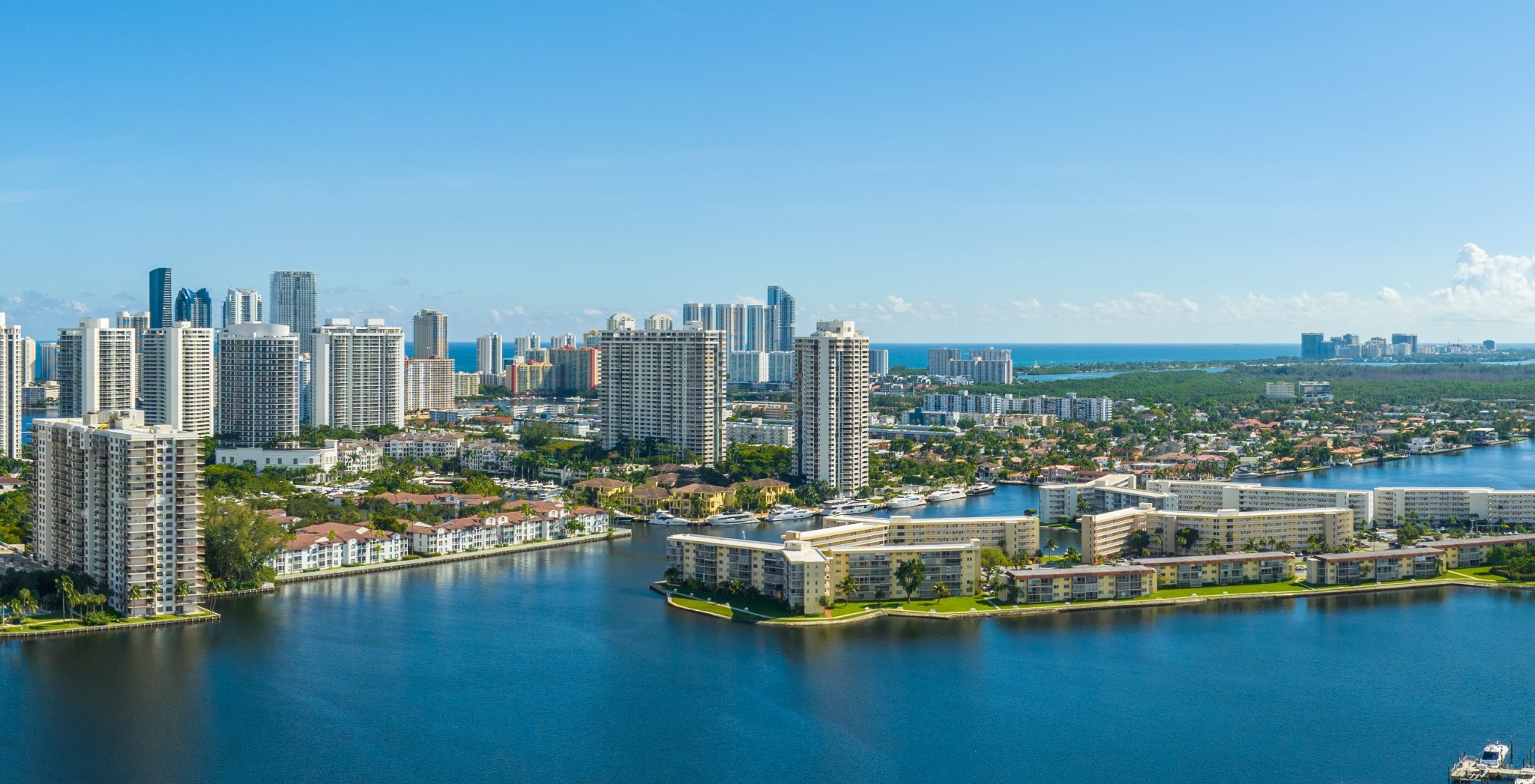 Aventura luxury and ultra luxury condos in an aerial waterfront view with high-rise residential towers, calm intracoastal canals, low-rise buildings, boats, and the ocean horizon under a clear blue sky.