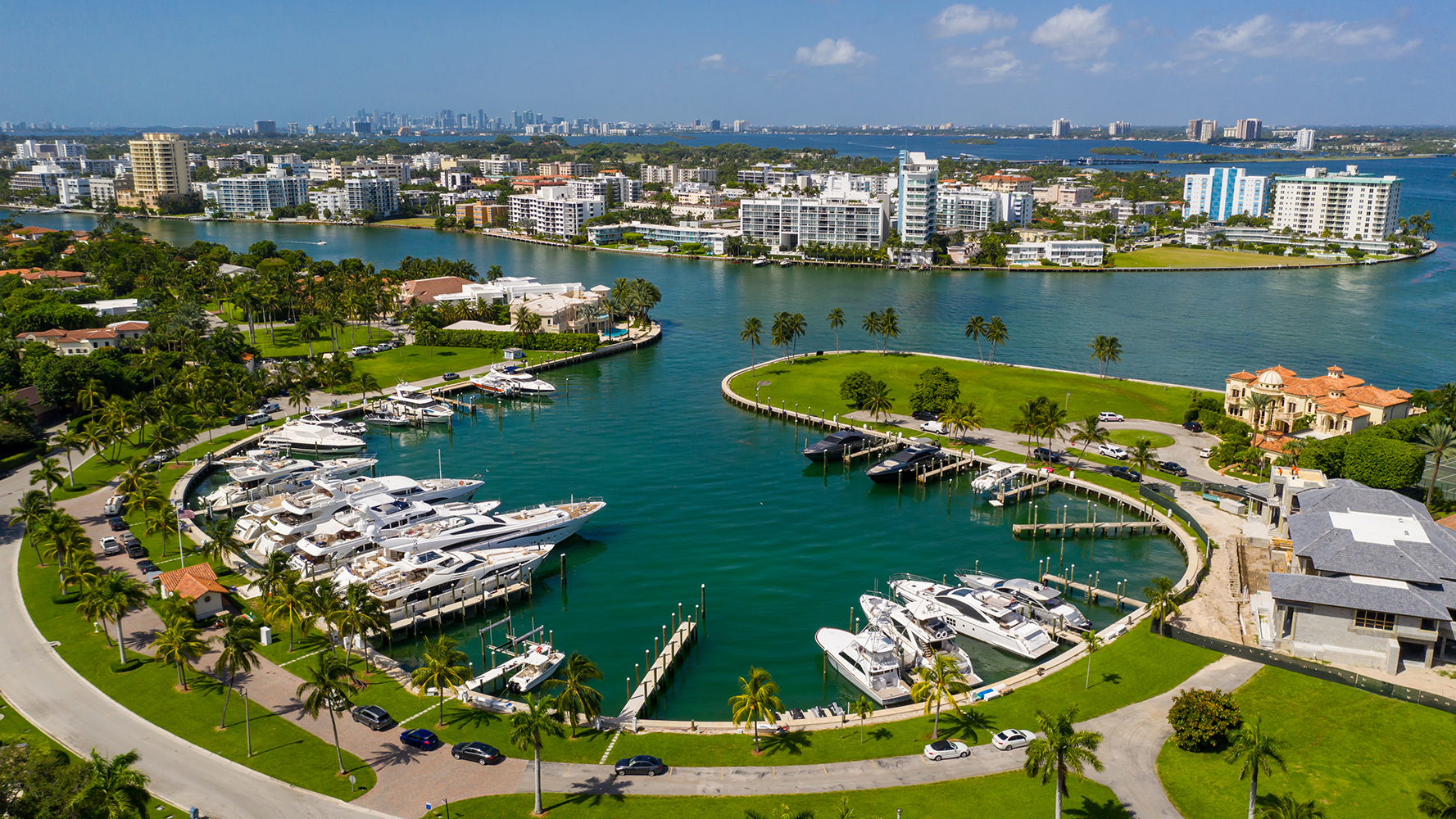 Aerial view of a marina with yachts, surrounded by water, palm trees, and modern buildings under a blue sky. Featuring Bal Harbour, waterfront, and Miami skyline.