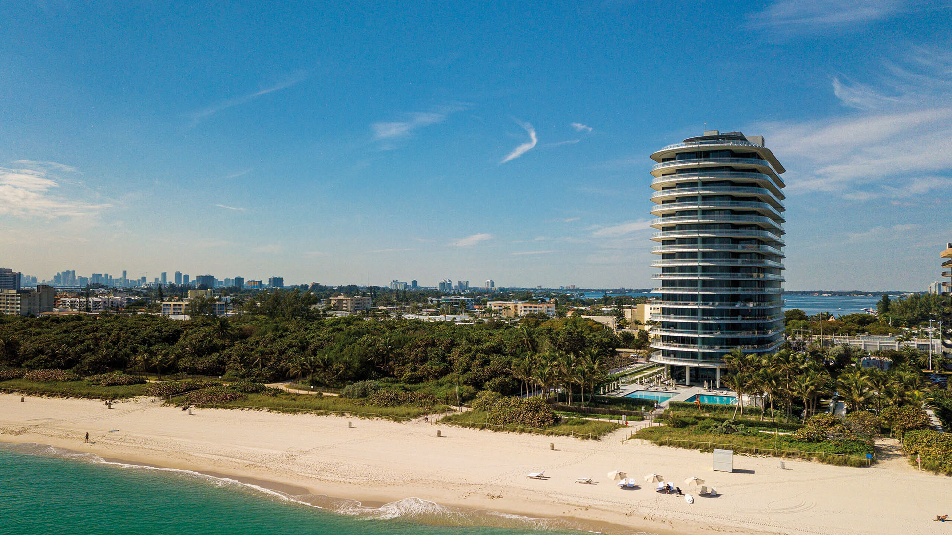 Eighty Seven Park, Miami Beach luxury and ultra luxury condos in a wide aerial beachfront view with sandy shoreline, tropical greenery, and the distant Miami skyline.