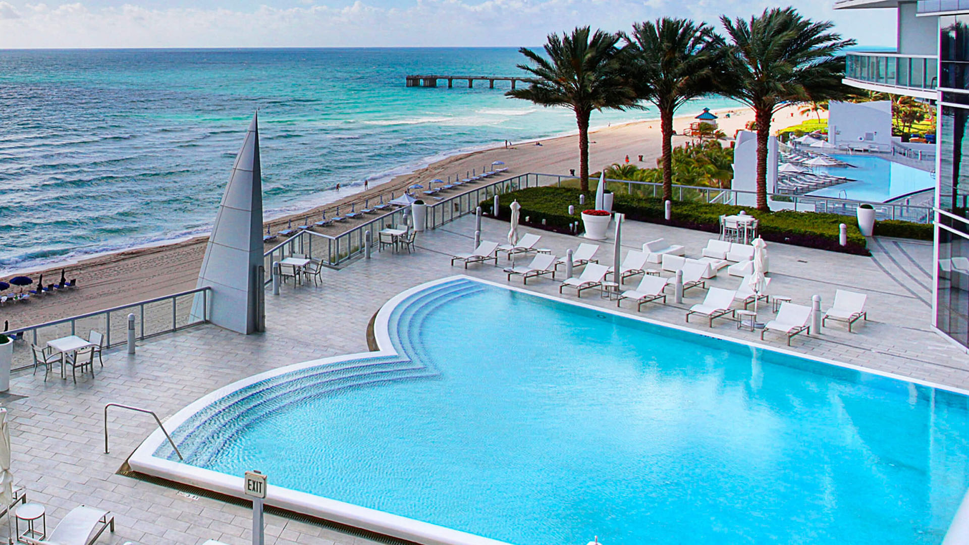 Beachfront pool terrace at Jade Ocean in Sunny Isles Beach, where luxury and ultra luxury condos overlook a curved oceanfront pool, palm-lined sundeck, white loungers, and the beach.
