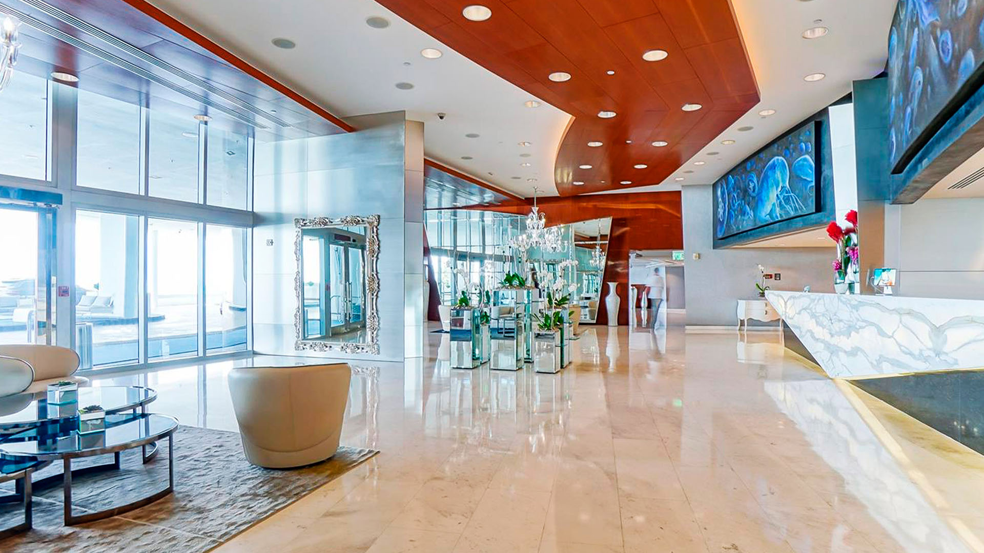 Grand lobby at Jade Ocean in Sunny Isles Beach for the luxury and ultra luxury condos, with a marble concierge desk, sculptural wood ceiling, mirrored finishes, and floor-to-ceiling glass.