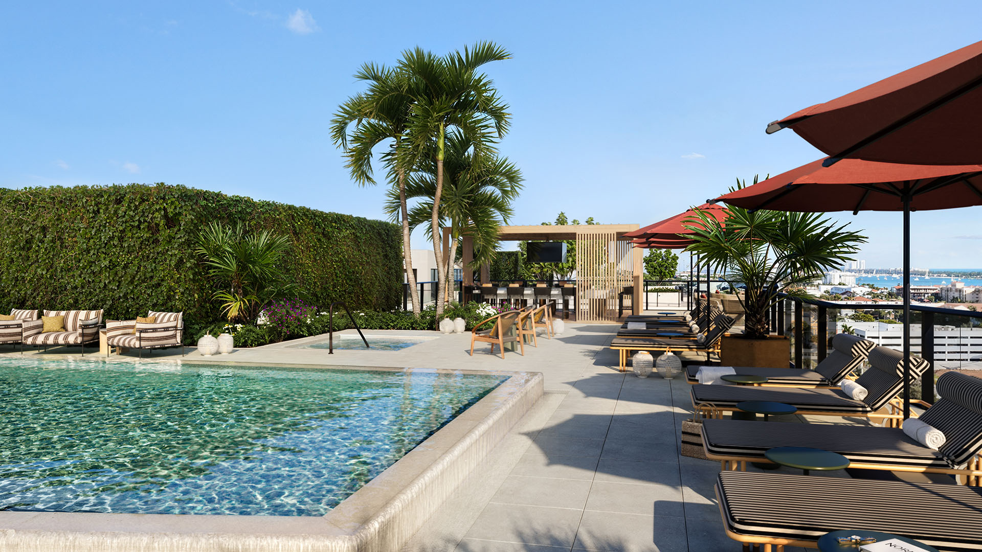 Rooftop pool deck at Nora House in West Palm Beach, luxury and ultra luxury condos with red umbrellas, striped loungers, a pergola lounge, tropical landscaping, and open water views beyond the terrace.